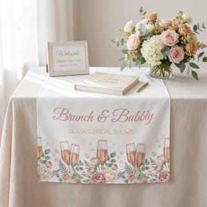 Pu&ograve; includere: Una tovaglia bianca con la scritta "Brunch & Bubbly" in rosa, progettata per un addio al nubilato. La tovaglia presenta calici di champagne e illustrazioni floreali. Un cartello incorniciato e un libro degli ospiti sono anche sul tavolo, insieme a una composizione floreale.