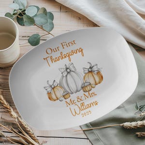 May include: White rectangular serving platter with rounded corners. The platter features the text "Our First Thanksgiving" and "Mr. & Mrs. Williams 2025" in gold script. It is decorated with watercolor pumpkins and acorns in shades of orange and gray.