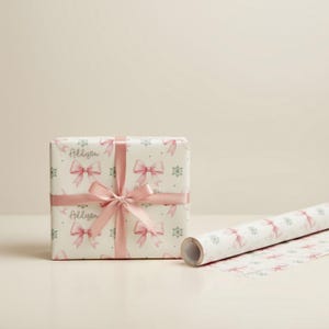 A square gift box wrapped in pink bow paper with a satin bow stands next to an unrolled tube of matching gift wrap.