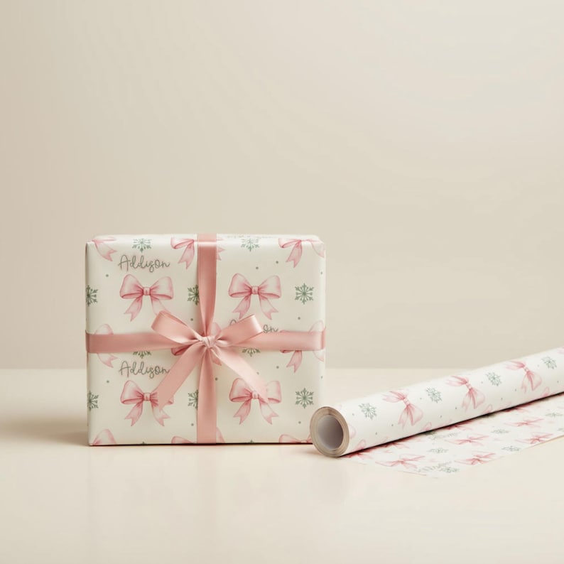 A square gift box wrapped in pink bow paper with a satin bow stands next to an unrolled tube of matching gift wrap.
