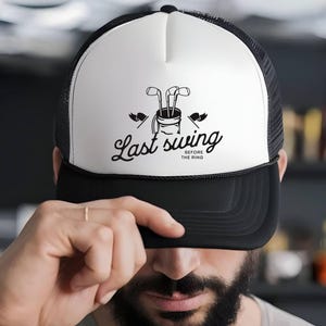 May include: A black and white trucker hat with a golf-themed graphic. The hat features a white front panel with a black illustration of golf clubs in a bag, and the text "Last swing before the ring". The brim and mesh back are black.