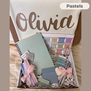 May include: A gift box filled with pastel-colored stationery and accessories. Includes a spiral-bound notebook, pens, sticky notes, and decorative clips. The box also contains a canvas bag with the name "Olivia" and the word "Pastels" printed on it.