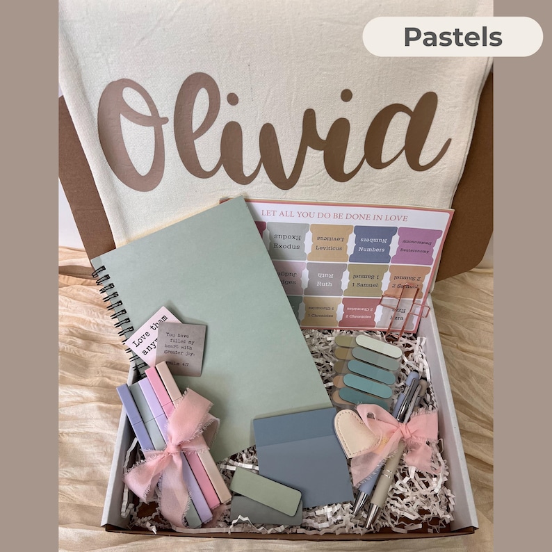 May include: A gift box filled with pastel-colored stationery and accessories. Includes a spiral-bound notebook, pens, sticky notes, and decorative clips. The box also contains a canvas bag with the name "Olivia" and the word "Pastels" printed on it.