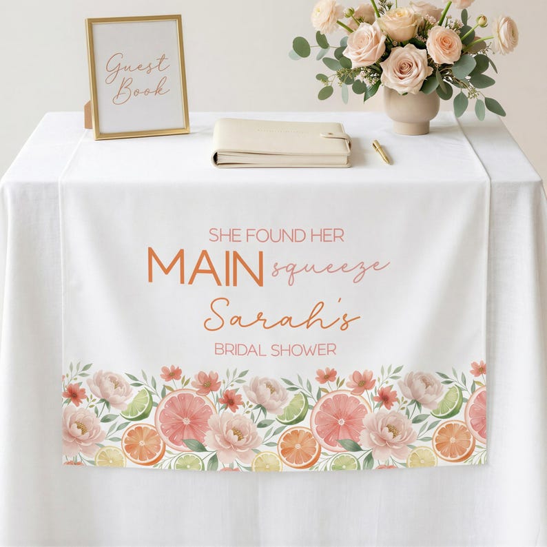 May include: White table runner with floral and citrus fruit border. Text reads "SHE FOUND HER MAIN squeeze Sarah's BRIDAL SHOWER." A gold framed sign says "Guest Book." A vase of flowers and a notebook are also on the table.