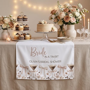 May include: A white table runner with the text "Bride in a tini bit OLIVIA'S BRIDAL SHOWER" in brown script. The runner is decorated with martini glasses, bows, and floral accents. The table is set with cupcakes, a cake, flowers, and candles.