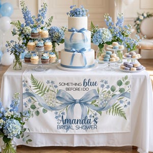 May include: A three-tiered white cake with blue watercolor accents and ribbon, surrounded by cupcakes, macarons, and floral arrangements. A table runner reads "Something Blue Before I Do" and "Amanda's Bridal Shower."