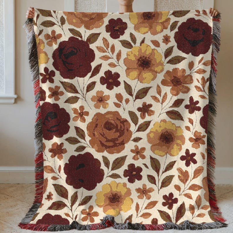 May include: A floral throw blanket featuring a cream background with a repeating pattern of large burgundy, brown, and yellow flowers, along with smaller flowers and leaves. The blanket has a fringed edge with red accents.