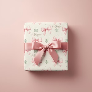 A square gift box wrapped in pink bow paper and tied with a wide satin ribbon rests on a pink surface.