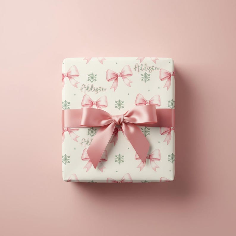 A square gift box wrapped in pink bow paper and tied with a wide satin ribbon rests on a pink surface.