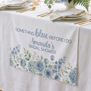 May include: White table runner with blue floral design and text that reads "SOMETHING blue BEFORE I DO Amanda's BRIDAL SHOWER". The runner is decorated with blue roses, hydrangeas, and other flowers. Gold silverware and plates are set on the table.