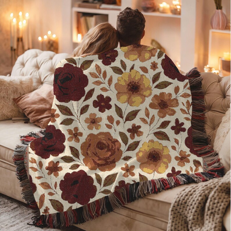 May include: Cream-colored throw blanket with a floral pattern in shades of burgundy, yellow, and brown. The blanket has a fringed edge and is draped over a couple sitting on a beige sofa.