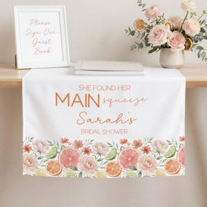 May include: White table runner with a floral and citrus design. The runner features the text "SHE FOUND HER MAIN squeeze Sarah's BRIDAL SHOWER" in orange script. The bottom border has watercolor illustrations of pink flowers, grapefruit, oranges, and limes.