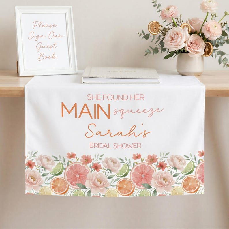 May include: White table runner with a floral and citrus design. The runner features the text "SHE FOUND HER MAIN squeeze Sarah's BRIDAL SHOWER" in orange script. The bottom border has watercolor illustrations of pink flowers, grapefruit, oranges, and limes.