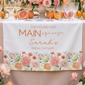 May include: White table runner with floral and citrus fruit border. Text reads "She found her MAIN squeeze Sarah's BRIDAL SHOWER". The design includes pink grapefruit, orange slices, lime, and flowers. Champagne glasses and fruit are on the table.