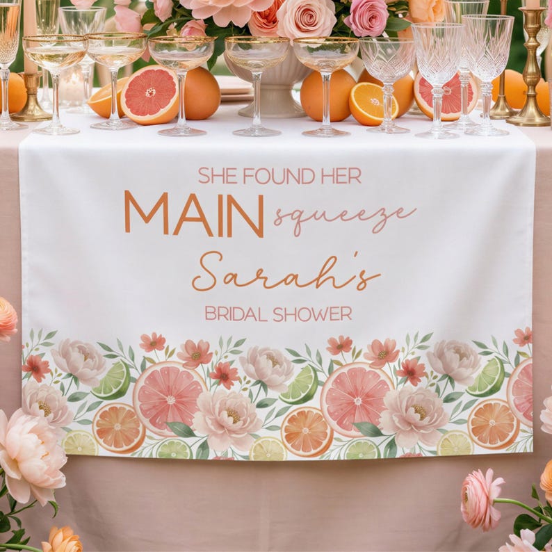 May include: White table runner with floral and citrus fruit border. Text reads "She found her MAIN squeeze Sarah's BRIDAL SHOWER". The design includes pink grapefruit, orange slices, lime, and flowers. Champagne glasses and fruit are on the table.