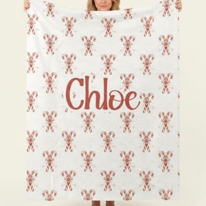 May include: A white blanket with a repeating pattern of red and white candy canes and bows. The name "Chloe" is printed in a large, red, cursive font in the center of the blanket. The blanket is held up by a person.