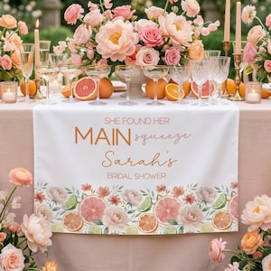 May include: A white table runner with the text "SHE FOUND HER MAIN squeeze Sarah's BRIDAL SHOWER" in copper-colored script. The runner is decorated with watercolor illustrations of citrus fruits and flowers. Champagne glasses and floral arrangements are on the table.