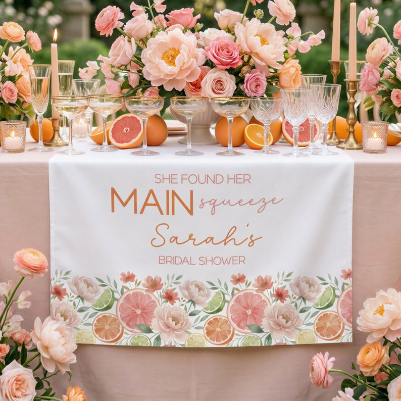 May include: A white table runner with the text "SHE FOUND HER MAIN squeeze Sarah's BRIDAL SHOWER" in copper-colored script. The runner is decorated with watercolor illustrations of citrus fruits and flowers. Champagne glasses and floral arrangements are on the table.