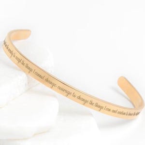 May include: A gold-colored cuff bracelet with engraved text. The bracelet has a curved design and is displayed on a white surface. The engraved text reads: "Serenity to accept the things I cannot change, courage to change the things I can, and wisdom to know the difference."