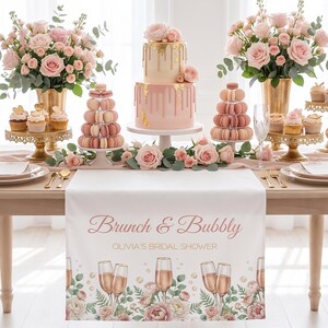 Pu&ograve; includere: Un'impostazione della tavola splendidamente decorata per un addio al nubilato. Il centrotavola &egrave; una torta rosa a due livelli con accenti dorati, circondata da rose rosa, cupcakes e macarons. Un runner da tavolo bianco mostra il testo "Brunch & Bubbly".