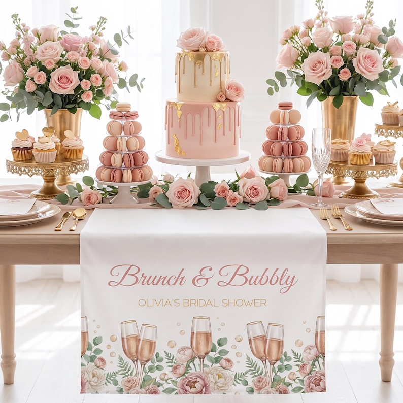 Pu&ograve; includere: Un'impostazione della tavola splendidamente decorata per un addio al nubilato. Il centrotavola &egrave; una torta rosa a due livelli con accenti dorati, circondata da rose rosa, cupcakes e macarons. Un runner da tavolo bianco mostra il testo "Brunch & Bubbly".