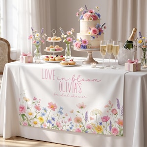 May include: A bridal shower table setting with a white tablecloth featuring a floral border and the text "LOVE in bloom OLIVIA'S bridal shower." The table includes a two-tiered cake, champagne, and various desserts.