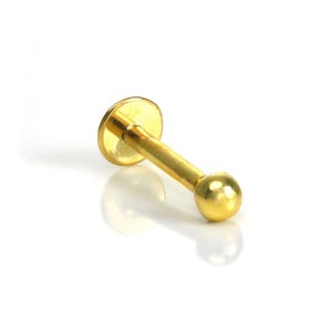 Puede incluir: Un piercing labial de color dorado. El piercing presenta un disco plano en un extremo, una barra recta y una pequeña bola en el otro extremo. La joya está sobre un fondo blanco.