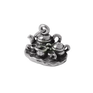 May include: A silver-toned metal charm featuring a miniature tea set on a scalloped tray. The set includes a teapot, a smaller lidded pot, and two small cups. A circular jump ring is attached to the top of the teapot for hanging purposes.