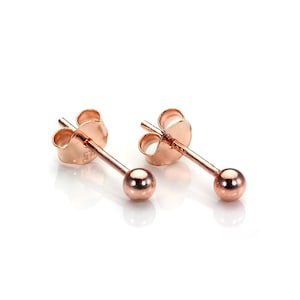 Op de afbeelding: Een paar roségouden bolvormige oorstekers. Elke oorbel heeft een kleine, ronde bal aan het einde van een slanke pin, met een vlindersluiting. De oorbellen worden gepresenteerd op een wit oppervlak, dat het licht reflecteert.