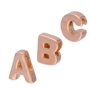 Op de afbeelding: Drie individuele bedels van roségoudkleurig metaal in de vorm van de letters A, B en C. Elke letterbedel heeft een glad, gepolijst oppervlak en een hol kanaal door het midden, ontworpen om aan een ketting of koord te worden geregen.