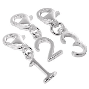 Puede incluir: Tres dijes de metal color plata con forma de los números uno, dos y tres. Cada dije está unido a un cierre de langosta mediante una pequeña argolla. Los dijes numéricos tienen el sello 925 grabado en el borde inferior de cada dígito.
