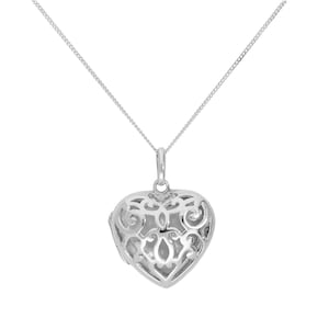 Puede incluir: Un collar con colgante en forma de corazón plateado. El colgante presenta un diseño calado abierto, revelando la forma interior del corazón. La cadena del collar es delicada y plateada, complementando los intrincados detalles del colgante. El collar se muestra sobre un fondo blanco.