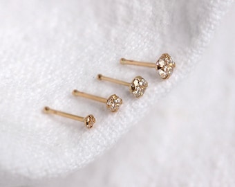 Könnte beinhalten: Vier goldene Nasenstecker mit klaren, funkelnden Edelsteinen. Die Stecker sind diagonal angeordnet, jeder mit einem unterschiedlich großen Edelstein. Die Goldstifte sind dünn und zart, vor einem weißen, strukturierten Hintergrund.