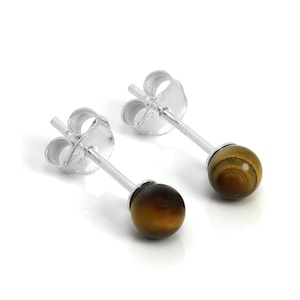 May include: A pair of stud earrings featuring a small, round, polished tiger's eye gemstone. The earrings have a silver post and backing. The tiger's eye stones display a mix of brown and gold colors. The earrings are set against a white background.