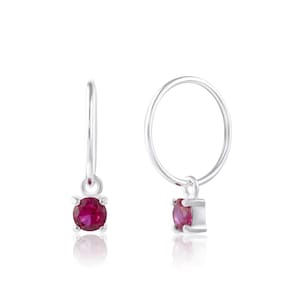 May include: A pair of silver hoop earrings, each with a round, deep red gemstone. The gemstones are set in a square silver setting, hanging from the bottom of the hoops. The earrings are shown against a white backdrop.