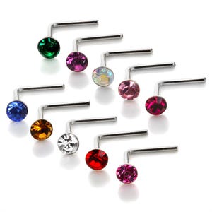 Puede incluir: Un surtido de piercings nasales en forma de L con piedras de cristal de varios colores. Los cristales incluyen verde, morado, iridiscente, rosa, rojo, azul, naranja, blanco y fucsia. El metal es plateado.