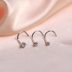 May include: Three silver nose studs, each featuring a clear, sparkling gemstone. The studs have a curved design with a small, round gem at the end. Set against a soft, pink fabric background.