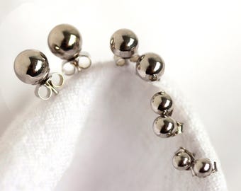 May include: Several pairs of silver-colored ball stud earrings are displayed on a white surface. The earrings have a shiny, reflective finish and are arranged in a curved pattern. The earrings are small and simple, with a classic design.