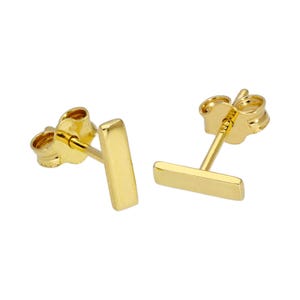 May include: A pair of gold-colored bar stud earrings. Each earring features a small, rectangular bar design attached to a post with a butterfly clasp. The earrings are presented on a white background.