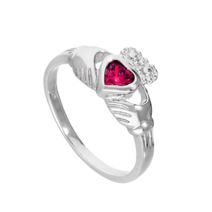 Peut inclure: Bague Claddagh en argent avec une pierre rouge rubis en forme de cœur. La conception de la bague comprend deux mains tenant le cœur, surmontées d'une couronne. L'anneau est sur fond blanc.