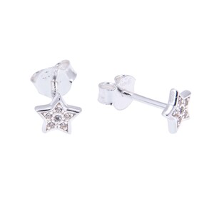 Puede incluir: Un par de pendientes de estrella de plata. Cada pendiente presenta un diseño de estrella de cinco puntas con pequeñas piedras preciosas transparentes en el centro. Se muestran sobre un fondo blanco.