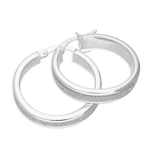 May include: A pair of silver hoop earrings. Each earring has a smooth, polished outer band and a textured, glittery inner band. The earrings are shown against a white background.