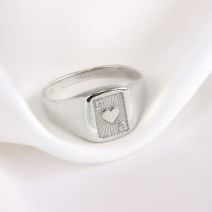 Puede incluir: Un anillo de sello plateado con una cara rectangular que presenta un corazón y las letras "r" y "a". El anillo tiene un acabado pulido y está sobre un fondo blanco.