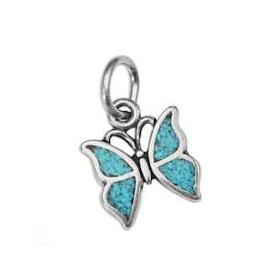 May include: A silver butterfly charm featuring inlaid turquoise stone fragments within the wings. The pendant includes a circular jump ring at the top for attachment, showcasing a detailed metal outline and textured blue stone inlay throughout.