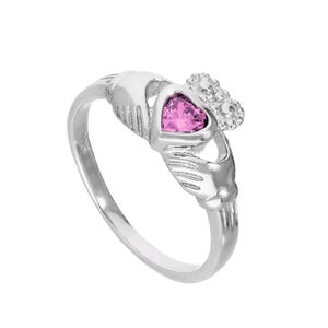 May include: A silver Claddagh ring with a pink heart-shaped gemstone. The ring features two hands holding a heart, and a crown. The ring is set against a white background.
