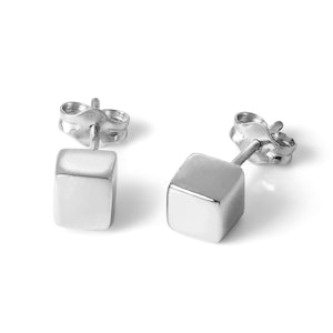 Könnte beinhalten: Ein Paar silberne Würfel-Ohrstecker. Jeder Ohrring hat einen kleinen, quadratischen, polierten Metallwürfel, der an einem Stecker mit einem Butterfly-Verschluss befestigt ist. Die Ohrringe sind auf weißem Hintergrund dargestellt.