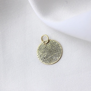 May include: A gold-colored, circular pendant featuring a detailed fingerprint design. The pendant has a small loop at the top for a chain. The background is a white, textured fabric.