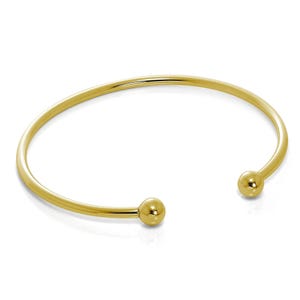 May include: A gold-coloured open cuff bracelet. The bracelet features a smooth, rounded band that curves into two small, spherical end caps. The bracelet is designed to slip over the wrist. The overall design is simple and elegant.