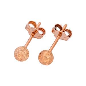May include: A pair of rose gold stud earrings. Each earring features a textured, spherical bead attached to a post with a butterfly backing. The earrings are displayed against a white background.
