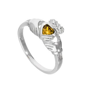 May include: A silver Claddagh ring with a heart-shaped yellow gemstone, clasped by hands and crowned. The ring, a symbol of love, loyalty, and friendship, is often given as a promise ring or wedding band.